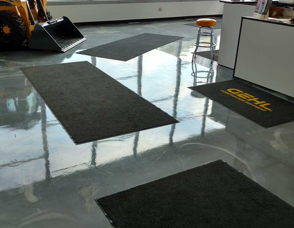 Polished Concrete Showroom Flooring with a glossy finish reflecting natural light, black mats, and a display of construction equipment and front desk area.